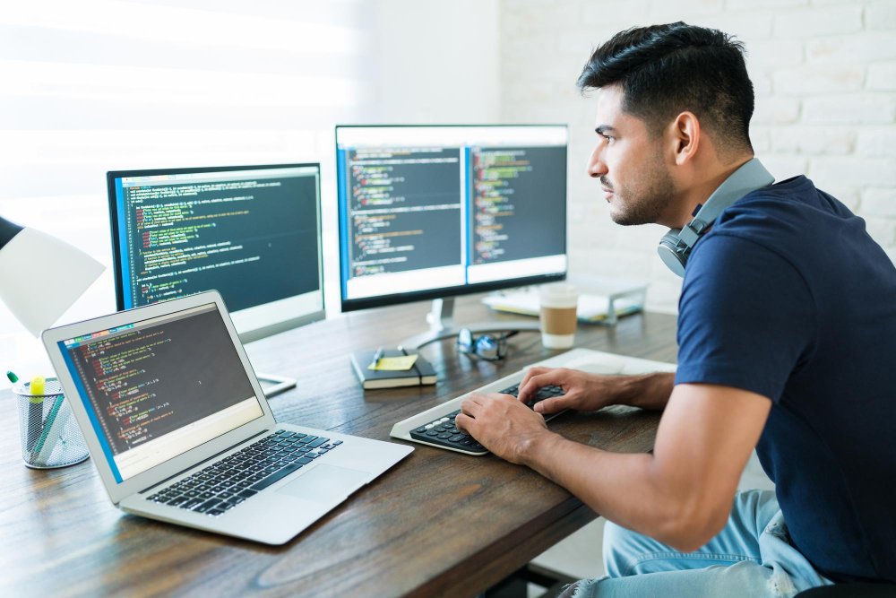 A focused male professional web developer coding on a laptop and two large desktop monitors with code visible on all screens. The developer is wearing a dark blue t-shirt and headphones around his neck, working at a clean wooden desk.